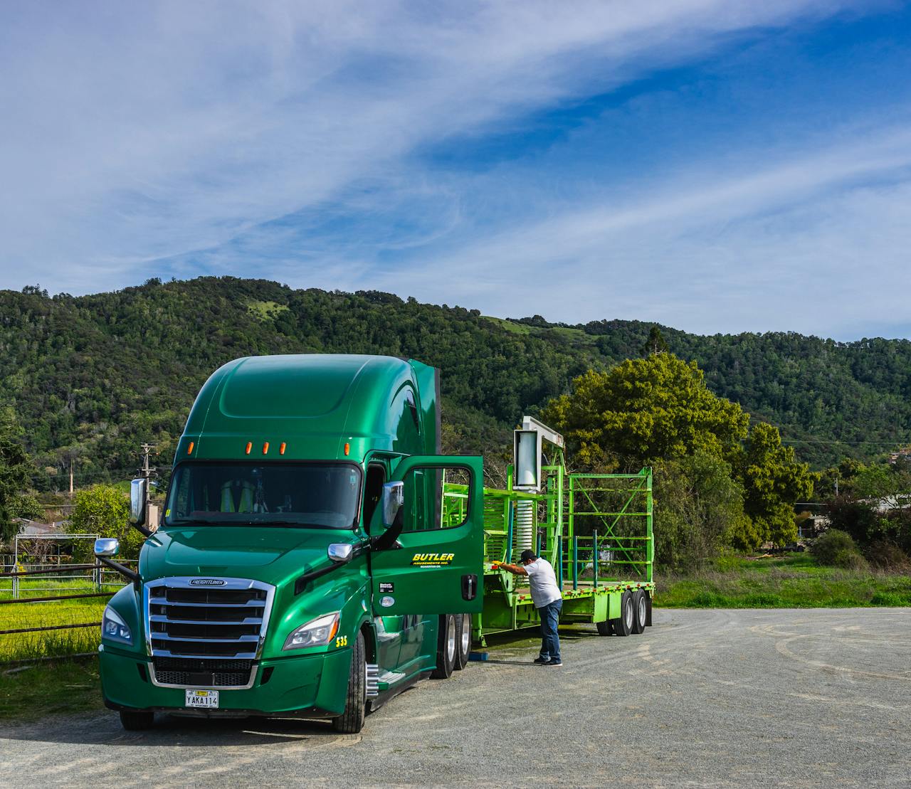 A vivid green truck with a trailer parked in a scenic landscape with hills and greenery.